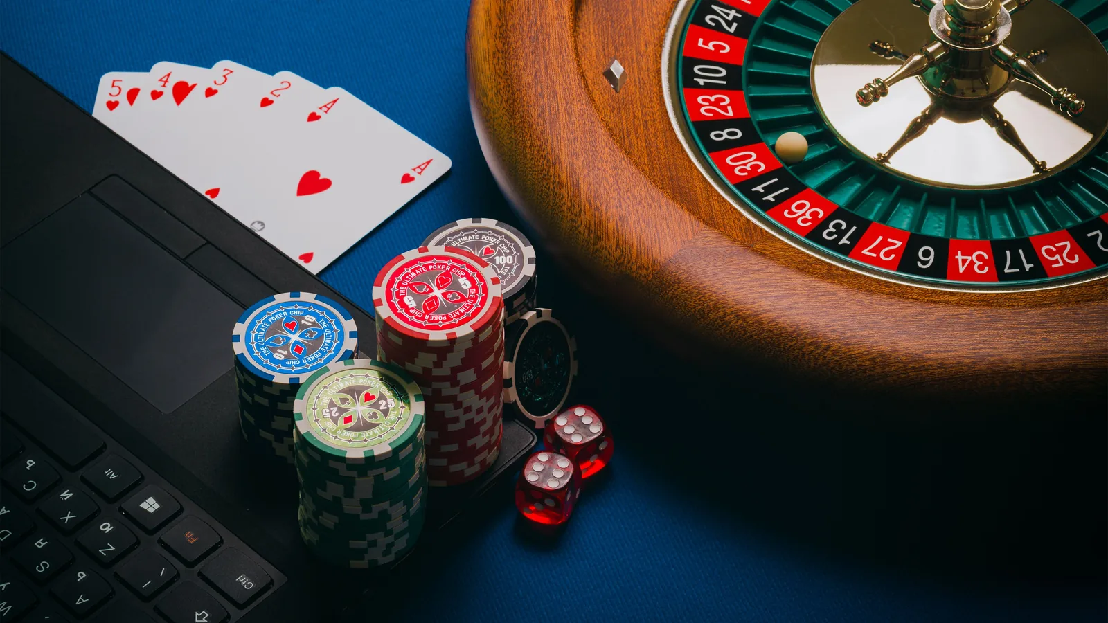 Laptop beside a roulette wheel, playing cards, casino chips and dice