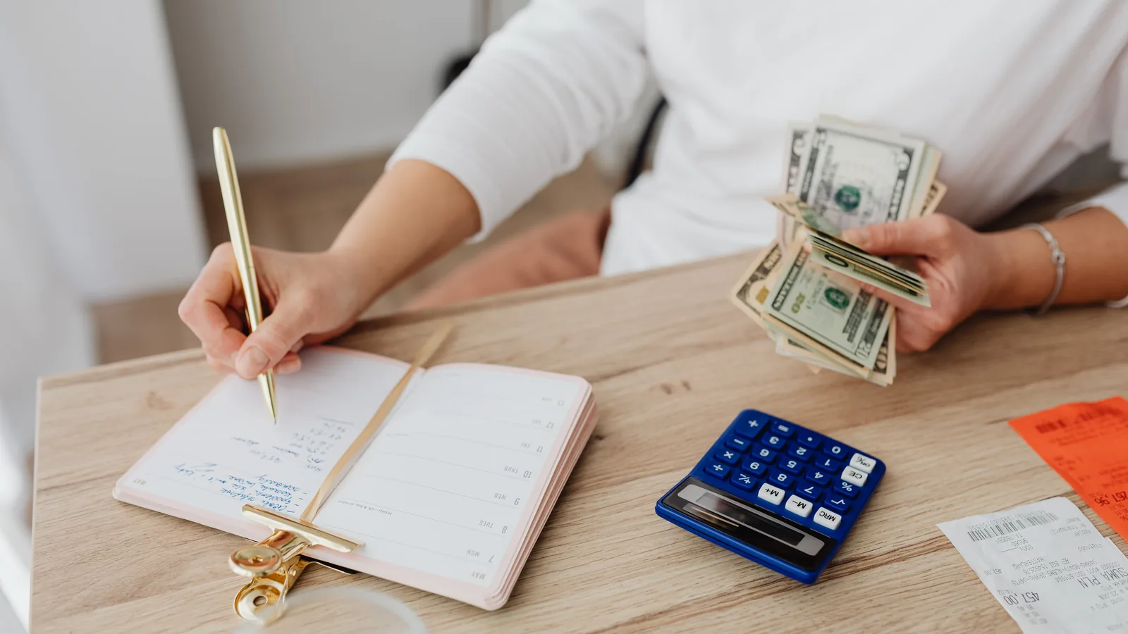 Person recording a budget beside cash, receipts and a calculator
